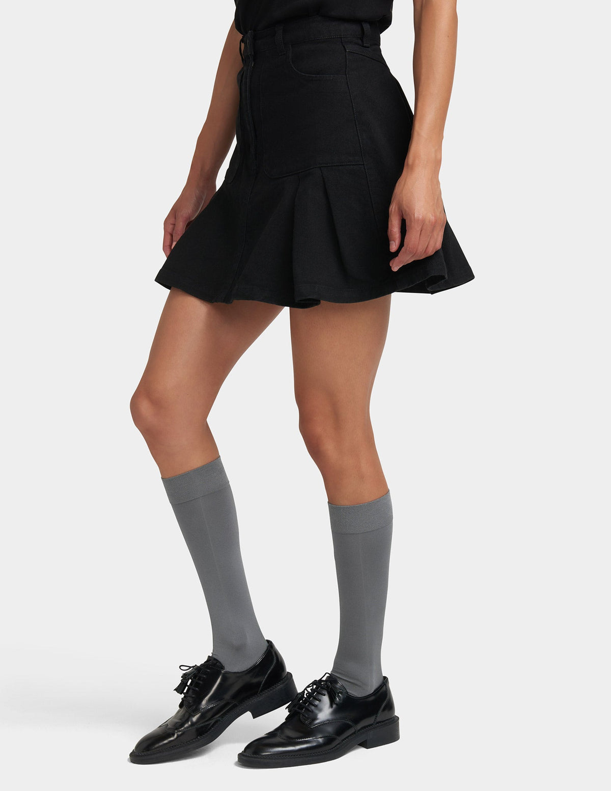 A model wearing a black pleated mini skirt, gray knee-high socks, and black leather oxford shoes, shown from the waist down against a light gray studio background.